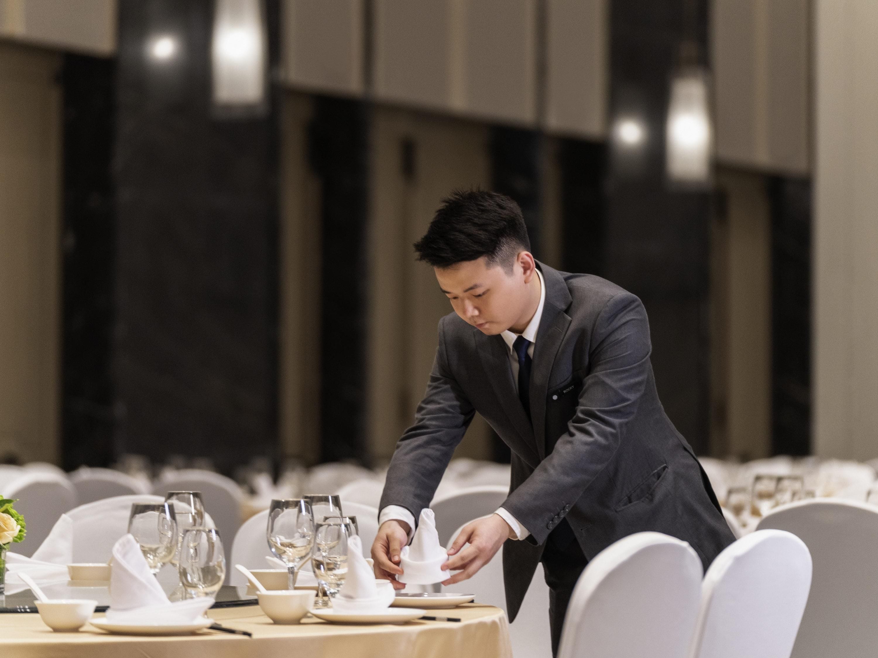 Un hombre preparando una mesa con manteles blancos y cristalería en un ambiente formal para cenar.