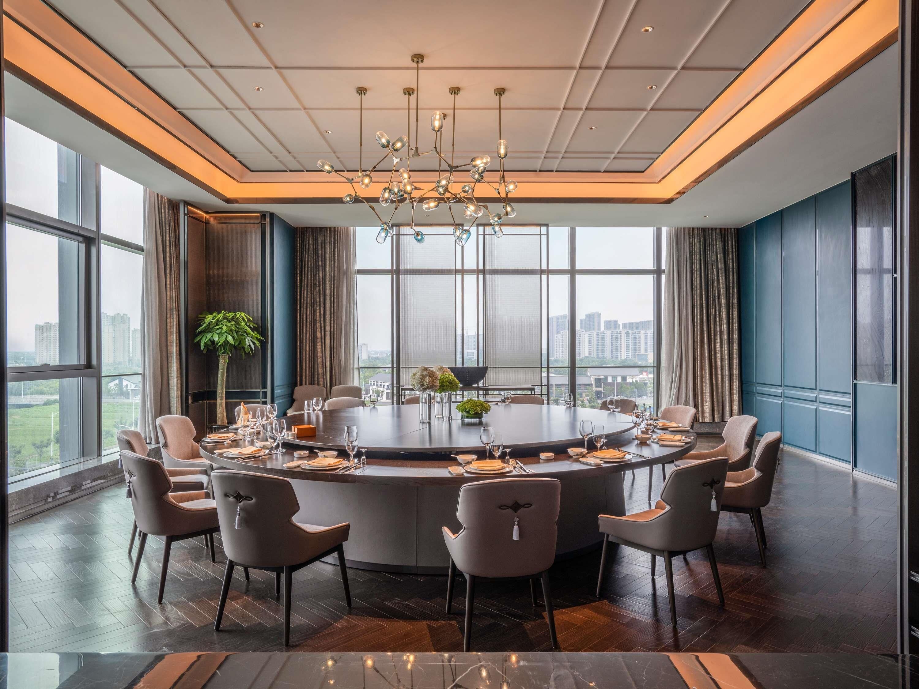 A modern conference room with a round table, multiple chairs, a chandelier, large windows, and a city view.