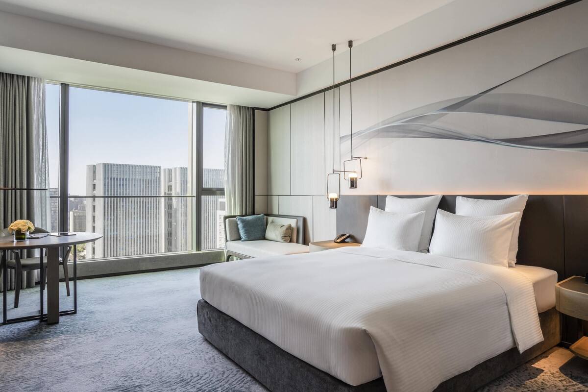 Une chambre d’hôtel moderne dotée d’un grand lit, d’un coin salon avec un fauteuil et une petite table, et offrant une vue sur la ville grâce à de grandes fenêtres.