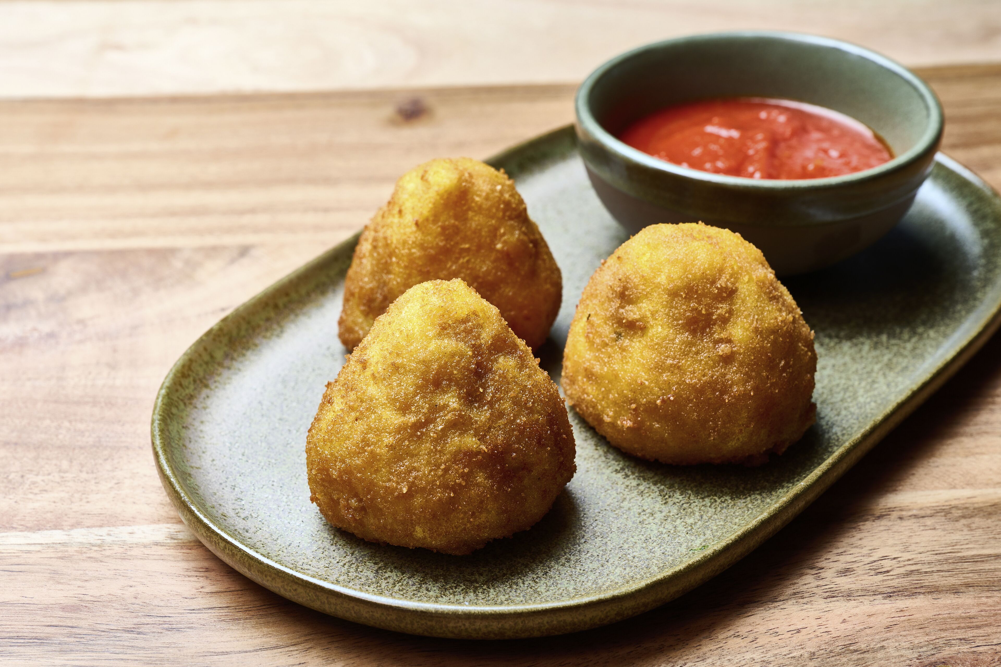 Three golden-brown croquettes on a green ceramic plate, accompanied by a small bowl of red sauce.