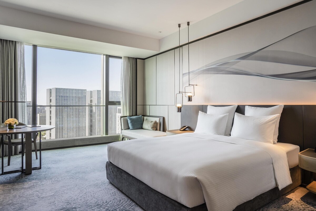 Una habitación de hotel moderna con una cama grande, un área de descanso con una silla, una mesita y vistas a la ciudad a través de amplios ventanales.