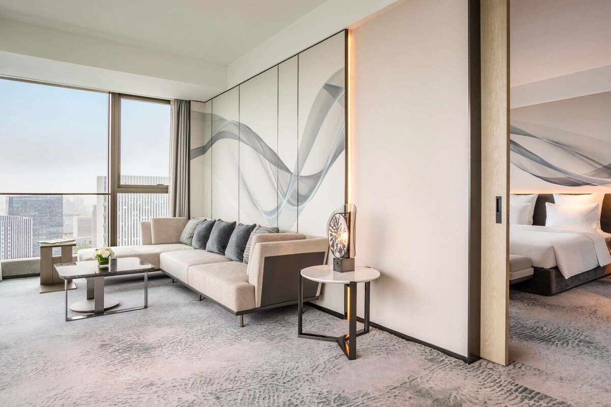 Una habitación de hotel moderna con una cama grande, un sofá, una mesita y un amplio ventanal con vistas a la ciudad.