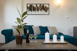 A living room with a teal sofa, a potted plant, a bottle of wine, two glasses, and three lit candles on a wooden table. - Alyvia Suites (Santorini)
