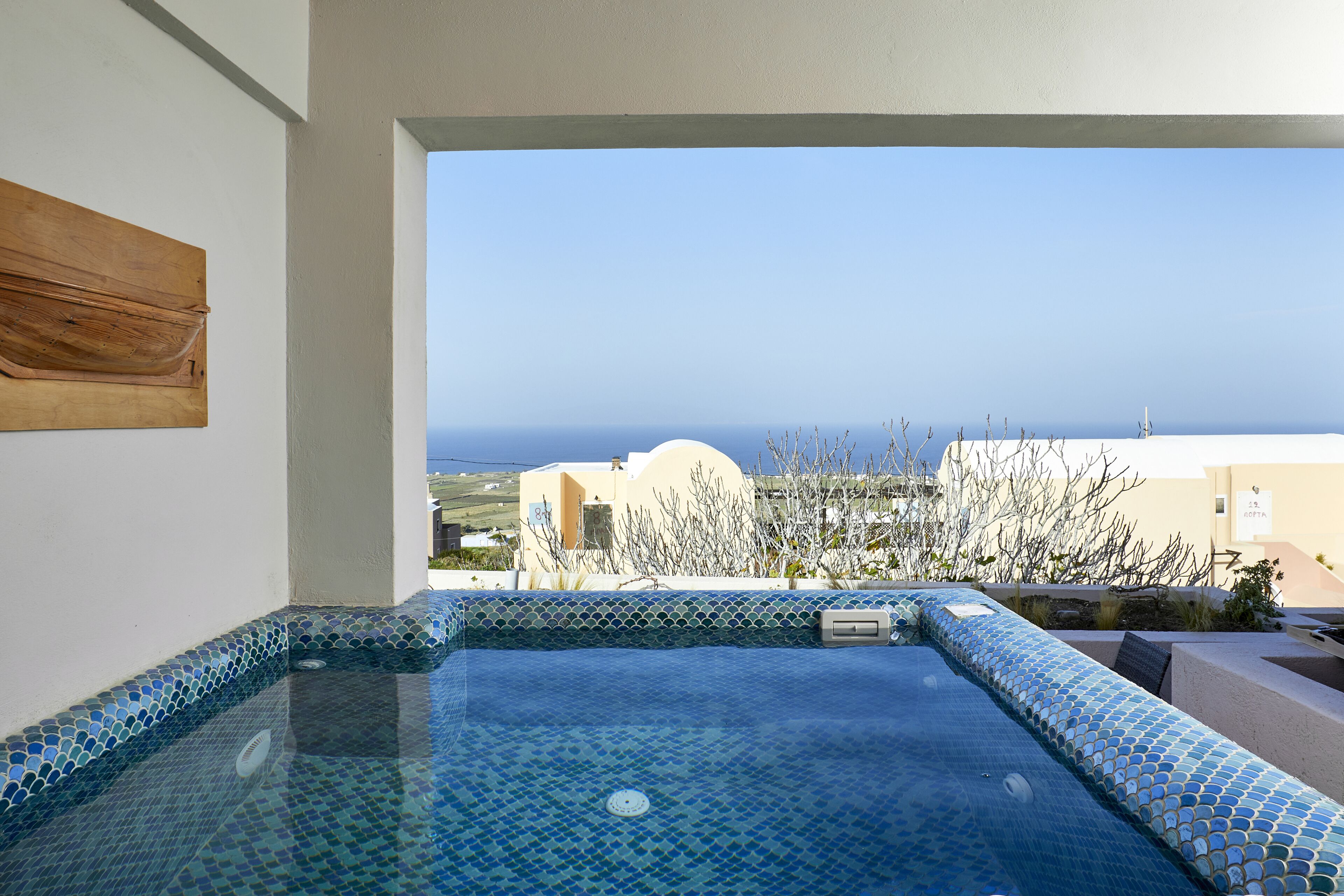 A clear view of a swimming pool with blue mosaic tiles, a wooden bench, and a view of the sea and buildings in the background.