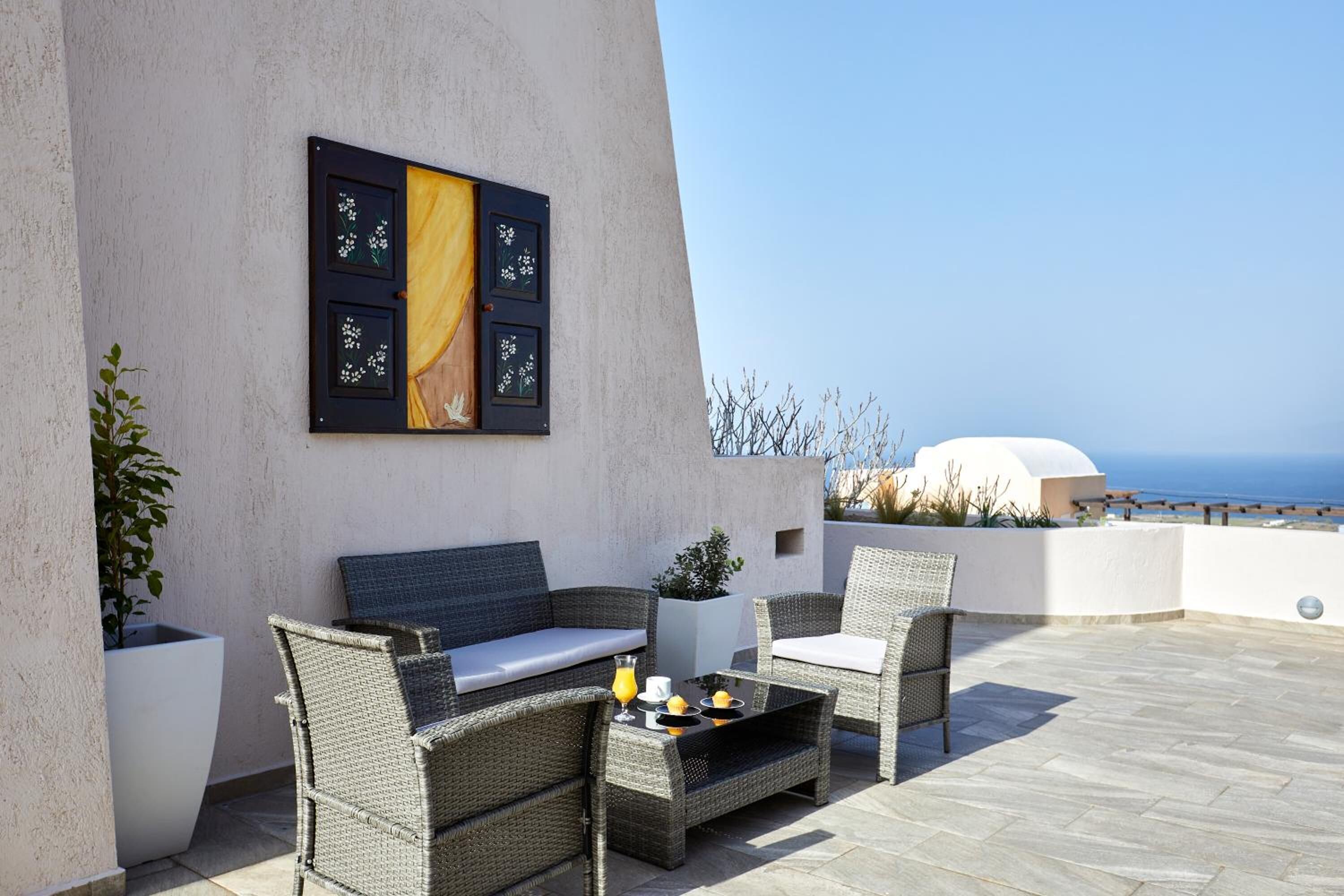 A terrace with wicker furniture, a white sofa, and a table with drinks. There is a view of the sea and a white dome structure in the background.