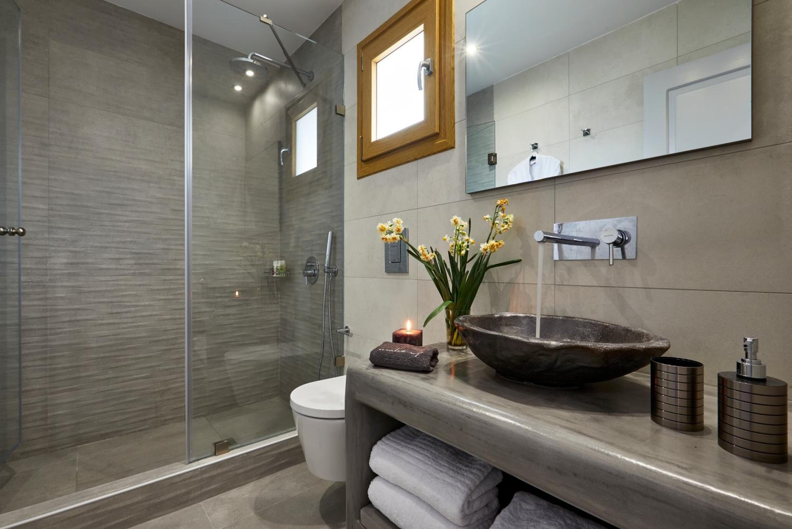A modern bathroom with a glass shower enclosure, a white toilet, a large mirror, a stone basin, and a small plant with yellow flowers.