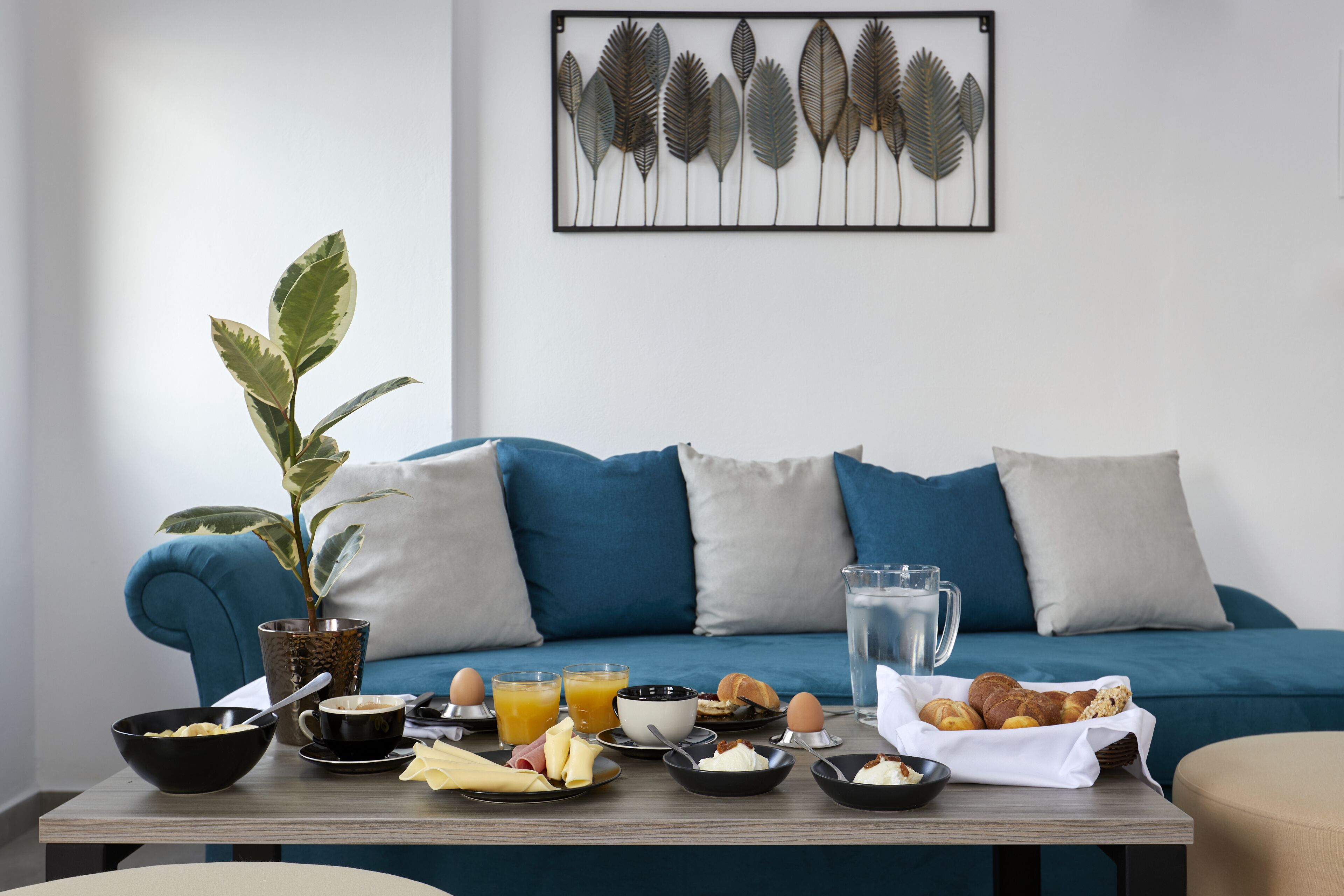 A breakfast setup on a table with a plant, a framed artwork, and a blue sofa in the background.