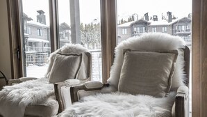 Deux fauteuils confortables, recouverts d’une étoffe moelleuse et agrémentés de plaids en fausse fourrure blanche, sont placés près de grandes fenêtres offrant une vue sur des immeubles d’habitation.