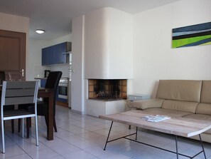 Uma sala de estar moderna com um sof á, uma mesa de centro e uma lareira. Há uma área de jantar com uma mesa e cadeiras ao fundo.