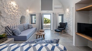 A modern room with a stone wall, a sofa with patterned cushions, a chair, a small table, and a TV on a wooden shelf. - Casa Tholari Al Mare (Αστυπάλαια)
