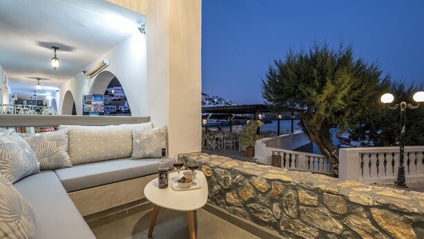 A balcony with a stone wall, a sofa, a small table with a bottle and snacks, and a view of a coastal town at dusk. - Casa Tholari Al Mare (Αστυπάλαια)