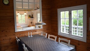 L'intérieur d'un chalet en bois comprend une table à manger, des chaises, un coin cuisine avec un poêle et une grande fenêtre donnant sur un paysage verdoyant.