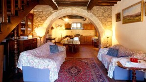 Un interior rústico con un arco de piedra que lleva a una cocina, dos sofás con fundas florales y una escalera de madera.
