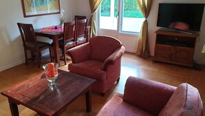 A living room with a wooden coffee table, red patterned sofa, dining table with chairs, and a TV on a wooden stand.