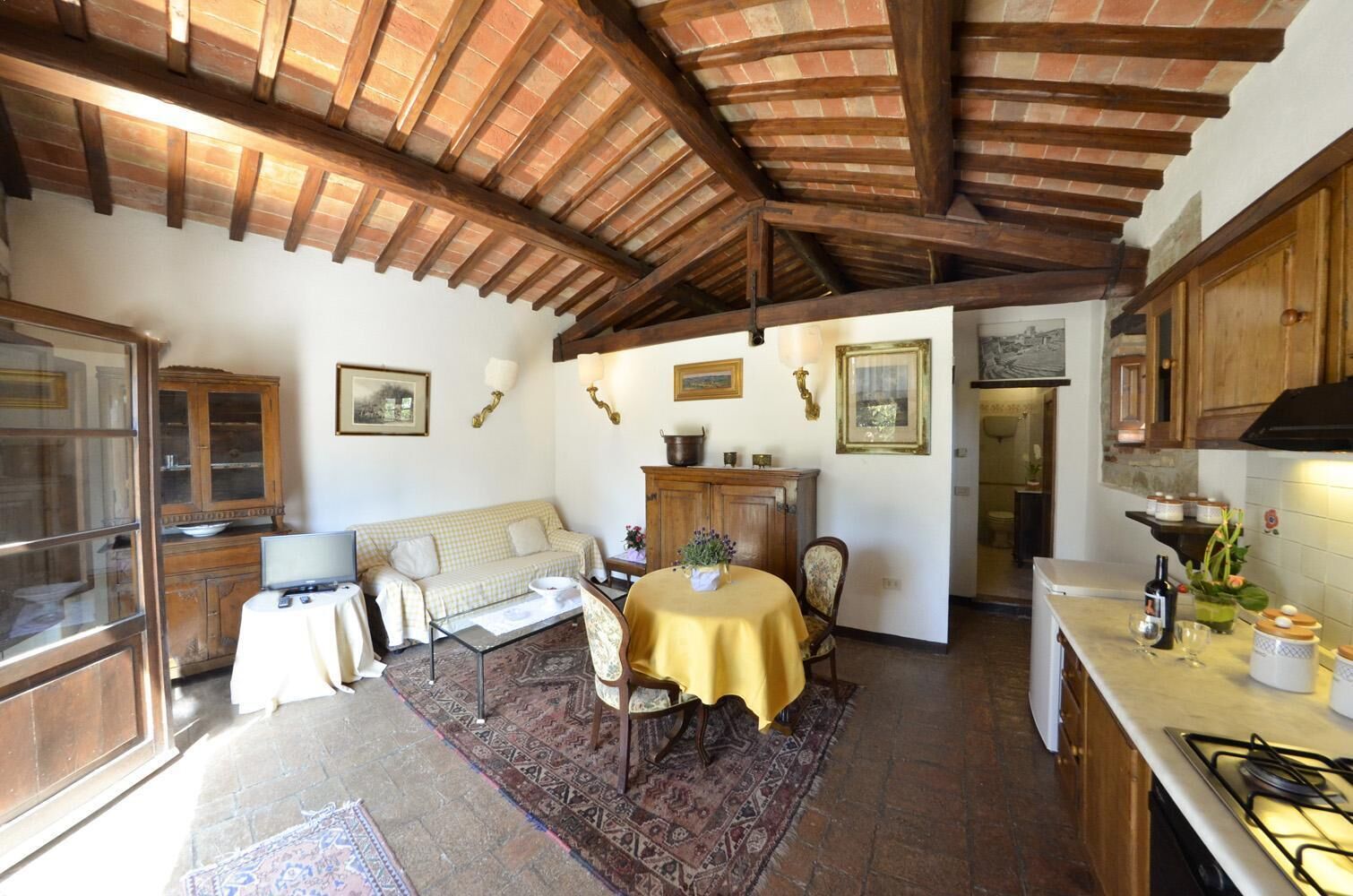 A rustic kitchen with exposed wooden beams, a dining area with a table and chairs, and a sofa in a living area.