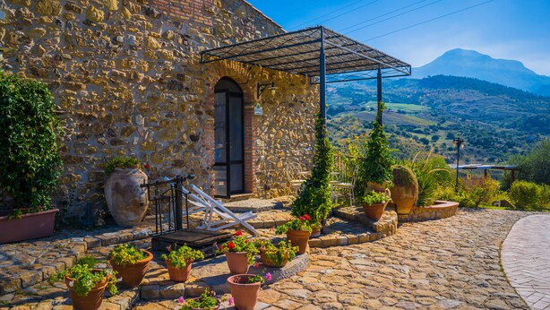 A stone building with a metal pergola, potted plants, and a cobblestone path. - Poggio Pozzetti (Lascari)
