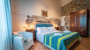 A bedroom with a stone wall, a bed with a striped blanket, a wooden wardrobe, and a nightstand with a lamp. - Poggio Pozzetti (Lascari)