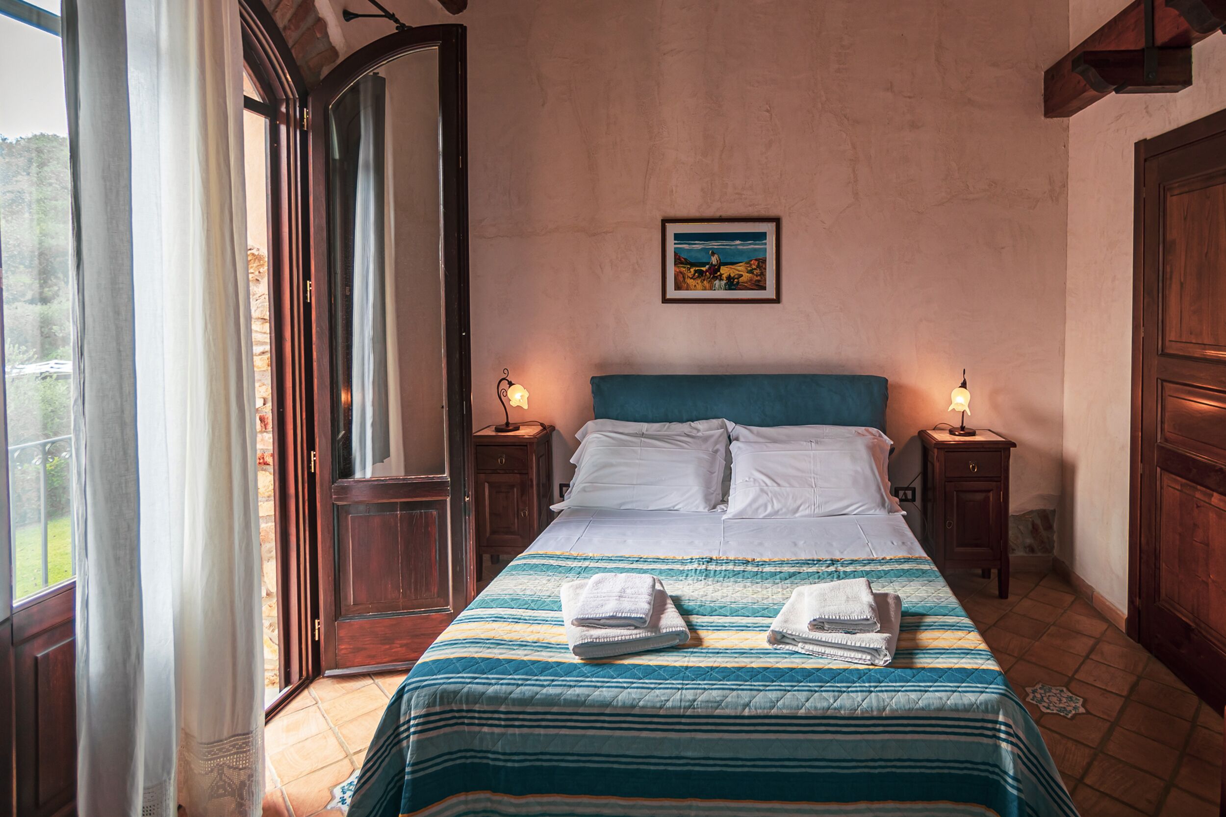 A bedroom with a bed, two bedside tables with lamps, a painting on the wall, and a large window with curtains.