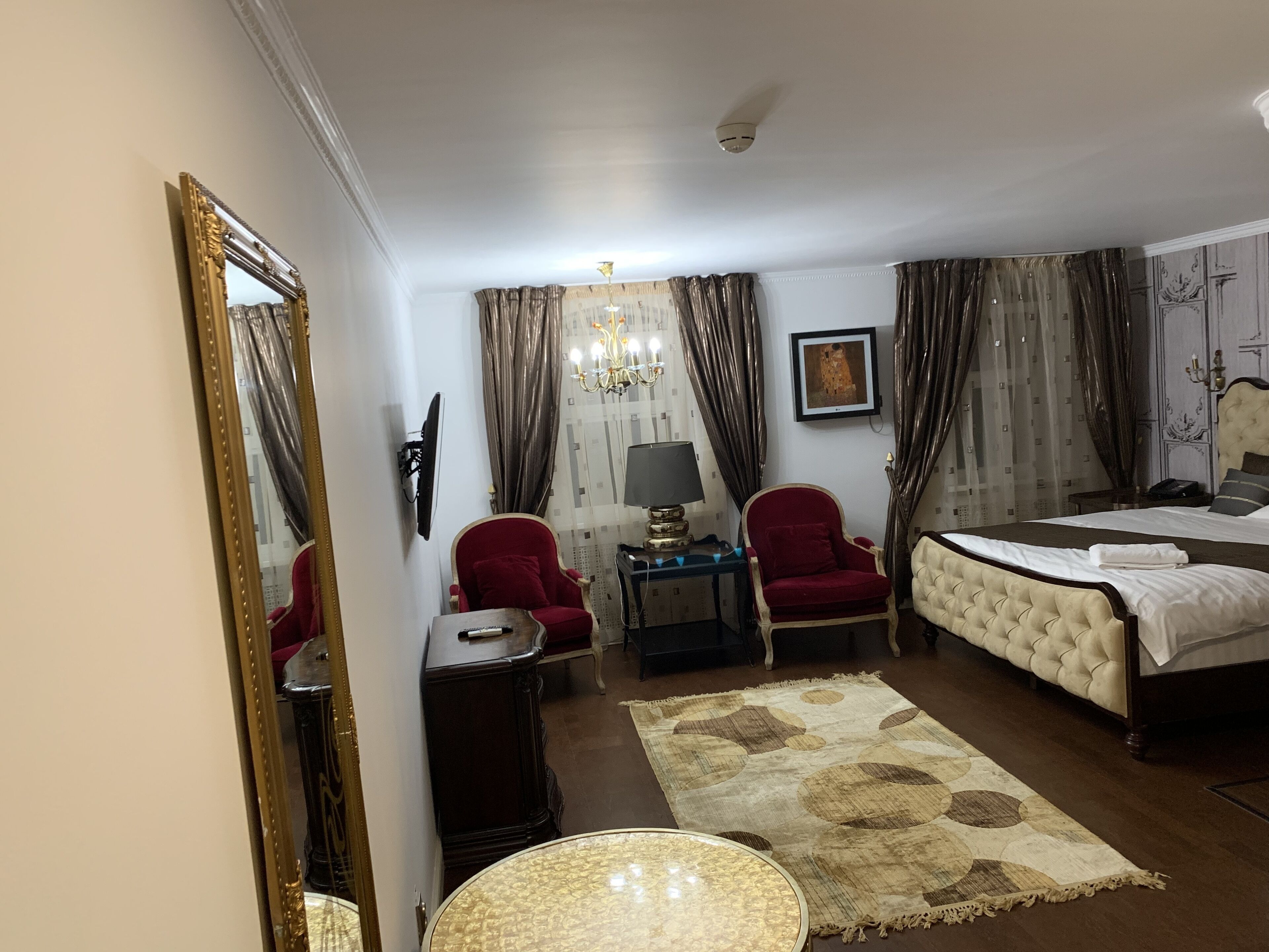 A hotel room with a bed, a red armchair, a small table with a lamp, a wall-mounted TV, and a large mirror.