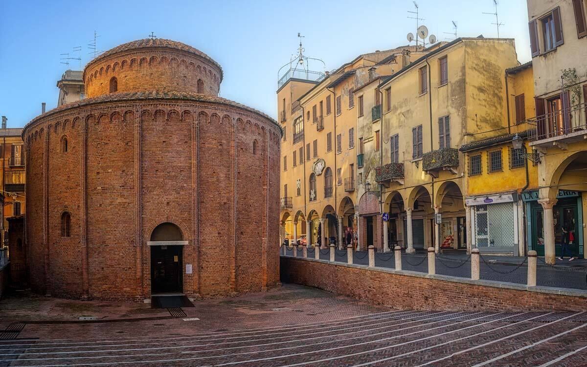 A historic brick building with a round tower, a courtyard with a paved area, and adjacent yellow buildings with balconies.