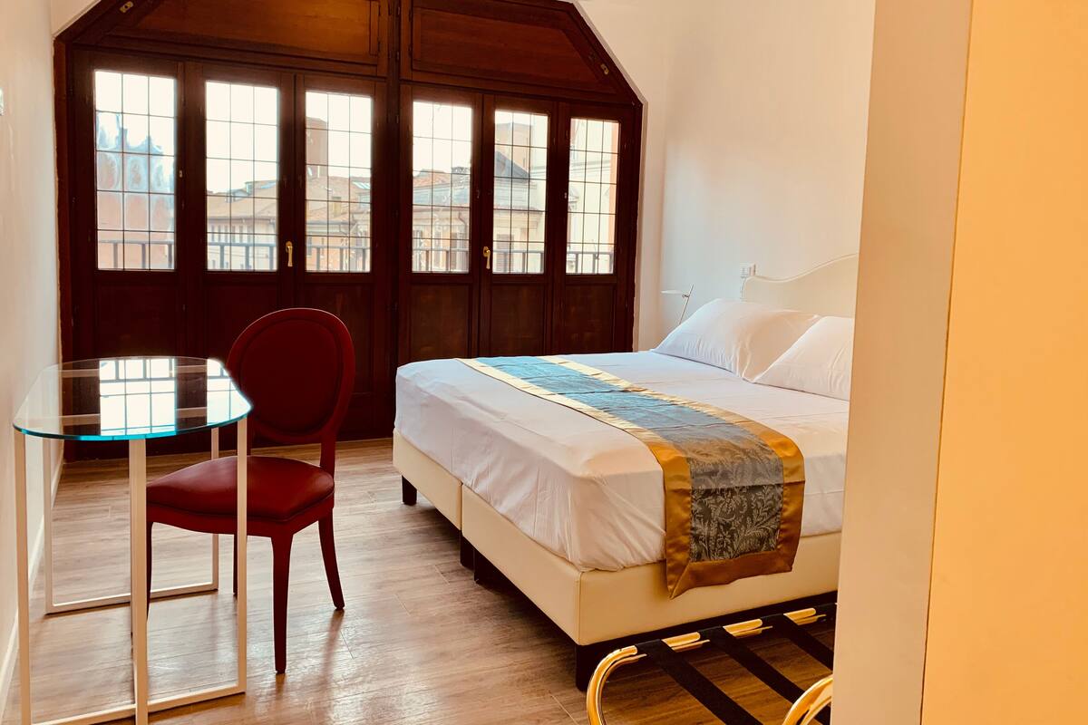 Ein Schlafzimmer mit einem Bett, einem roten Sessel, einem Glastisch und einem großen Fenster mit Blick auf Gebäude.