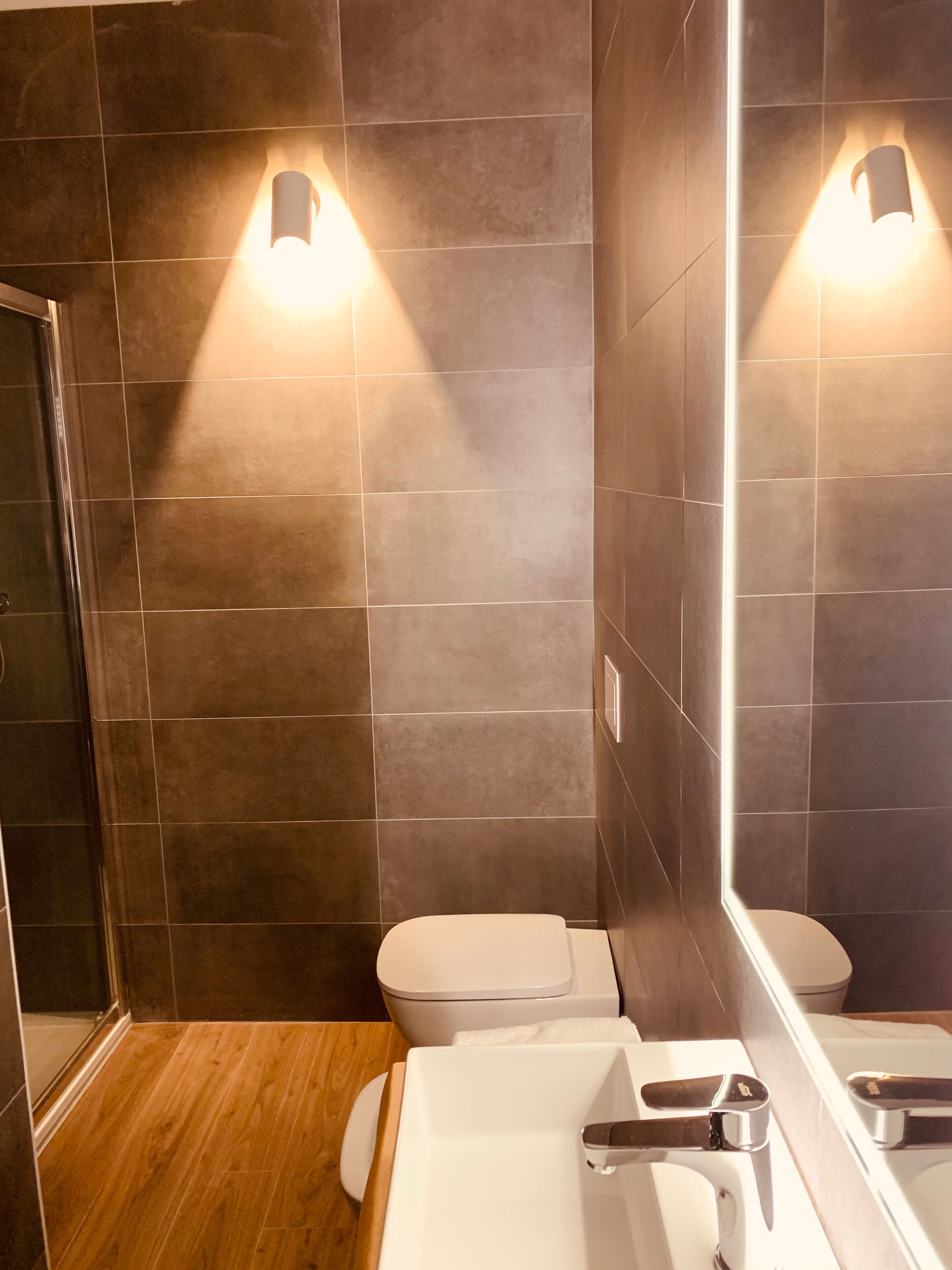 A modern bathroom with dark tiled walls, a wooden floor, a white toilet, and a white sink with a chrome faucet.