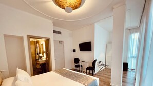 Habitació d'hotel amb un llit, un escriptori, una cadira, un televisor i un bany visible per una porta oberta.