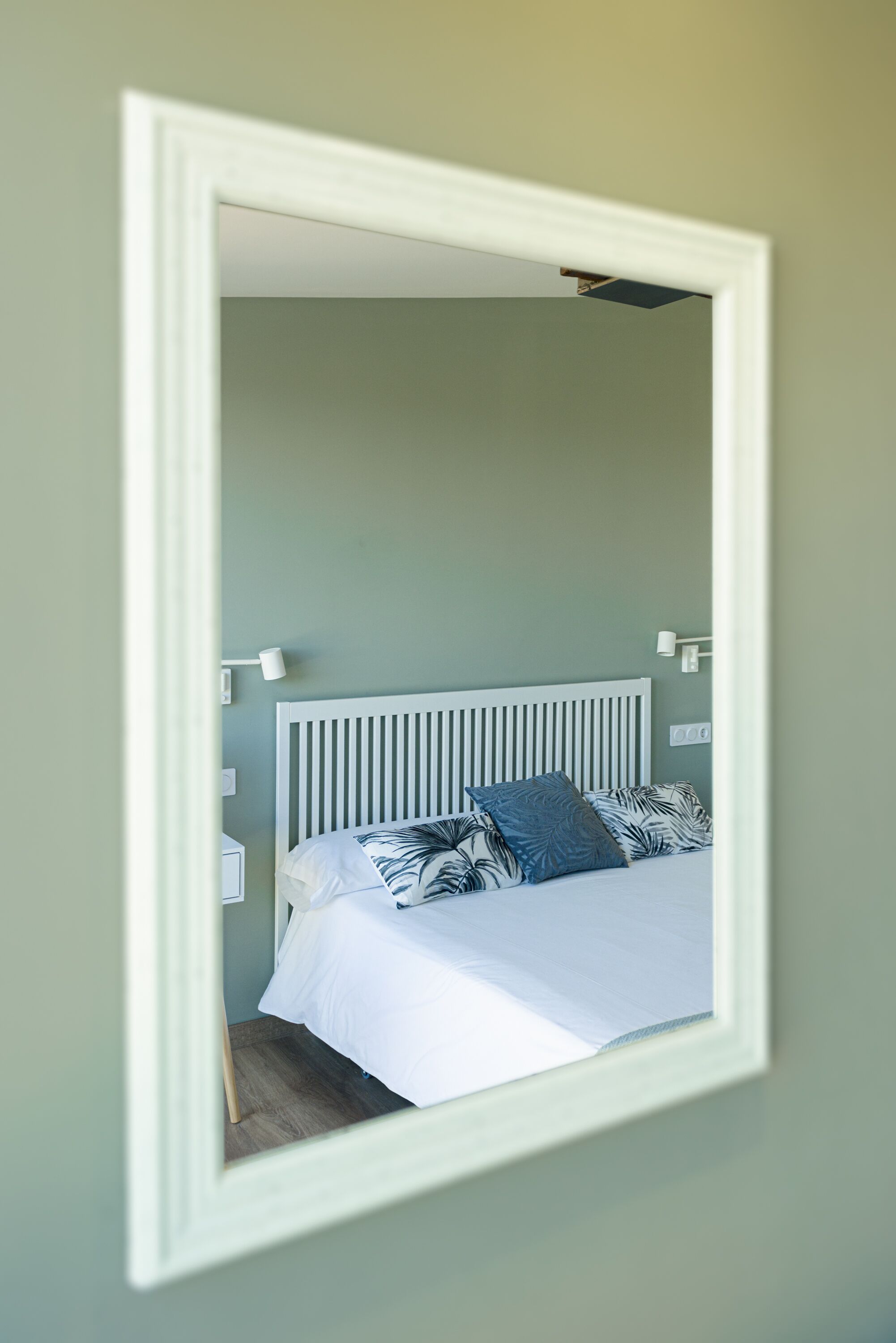 Un dormitorio con una cama blanca, cabecera blanca y espejo con marco blanco en la pared.