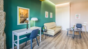 Una sala de estar moderna con paredes verde azulado, un escritorio blanco, una silla azul, un sofá color beige y una mesita redonda con una lámpara.
