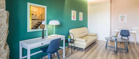 A modern living room with teal walls, a white desk, a blue chair, a beige sofa, and a small round table with a lamp.