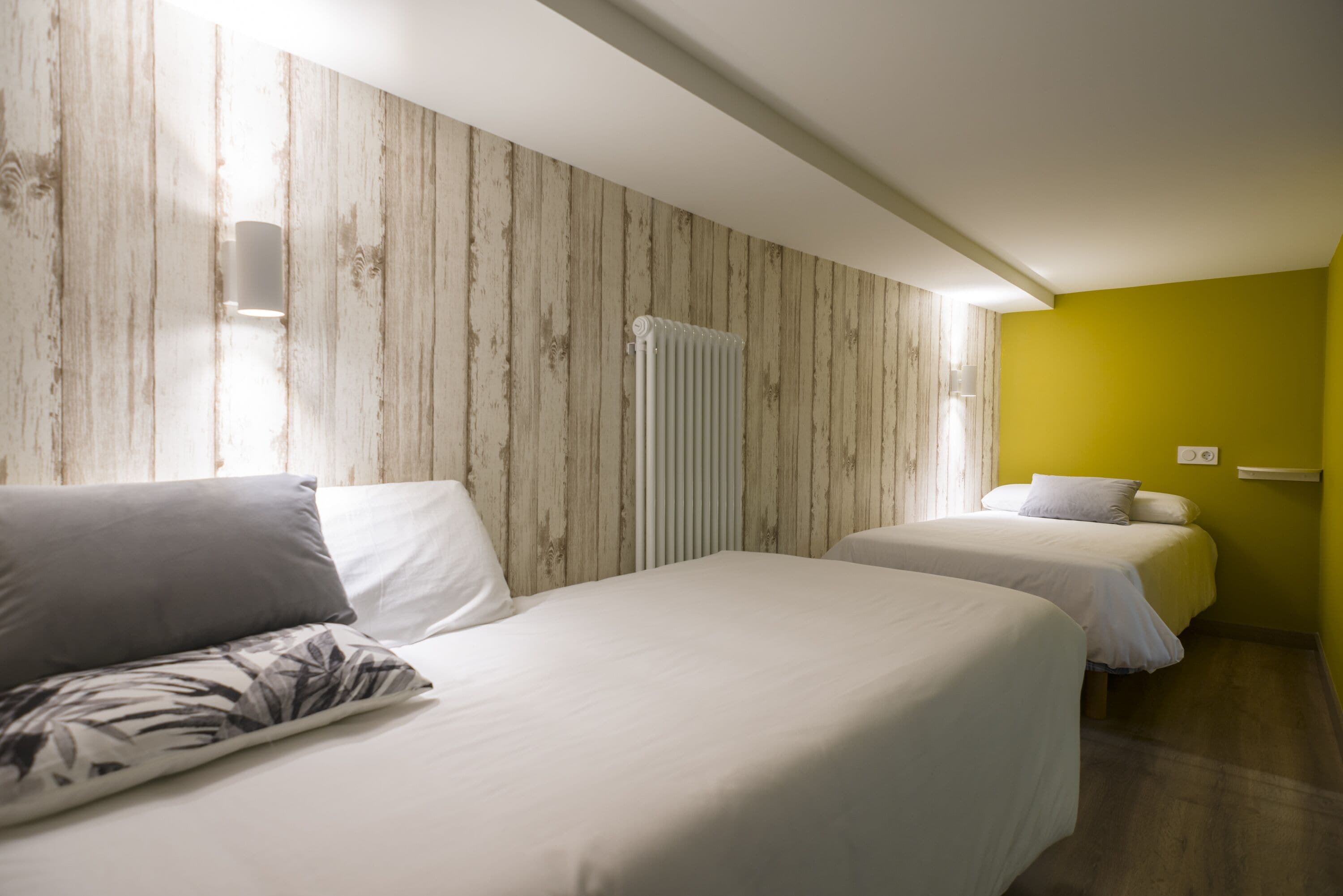 Quarto de hotel com duas camas, painéis de madeira nas paredes, radiador e uma parede com detalhe amarelo.