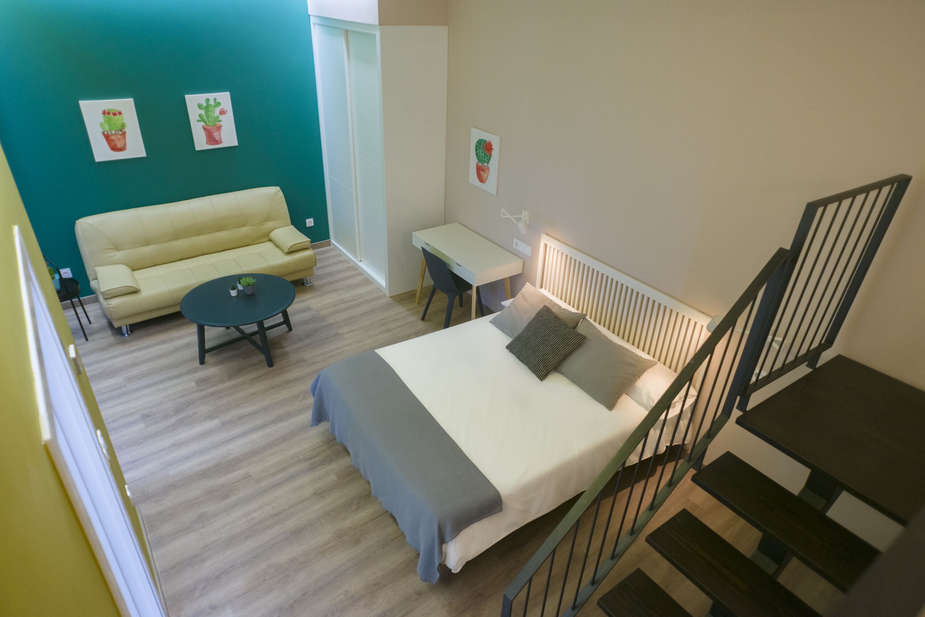 A bedroom with a bed, bedside table, and a desk with a chair. There is a sofa and a small table with a plant in the corner.