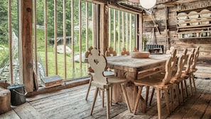 A rustic wooden dining area with a large table and chairs, a window with vertical blinds, and a shelf with glassware.