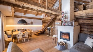 Ein rustikaler Innenraum mit einer Holztreppe, einem Kamin, einer Essecke mit Tisch und Stühlen sowie einer Sitzecke mit Sofa.