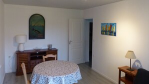 A dining area with a round table covered by a floral tablecloth, a wooden cabinet with a lamp, and a painting on the wall. - La Calypso the 2-person studio (+ 1 child under 3) ideal for fishing (Blangy-sur-Bresle)