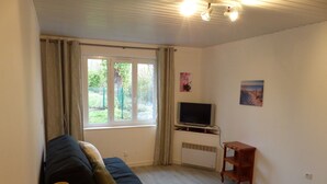 A living room with a dark-colored sofa, a small wooden side table, a television on a stand, a window with curtains, and a wall-mounted picture. - La Calypso the 2-person studio (+ 1 child under 3) ideal for fishing (Blangy-sur-Bresle)