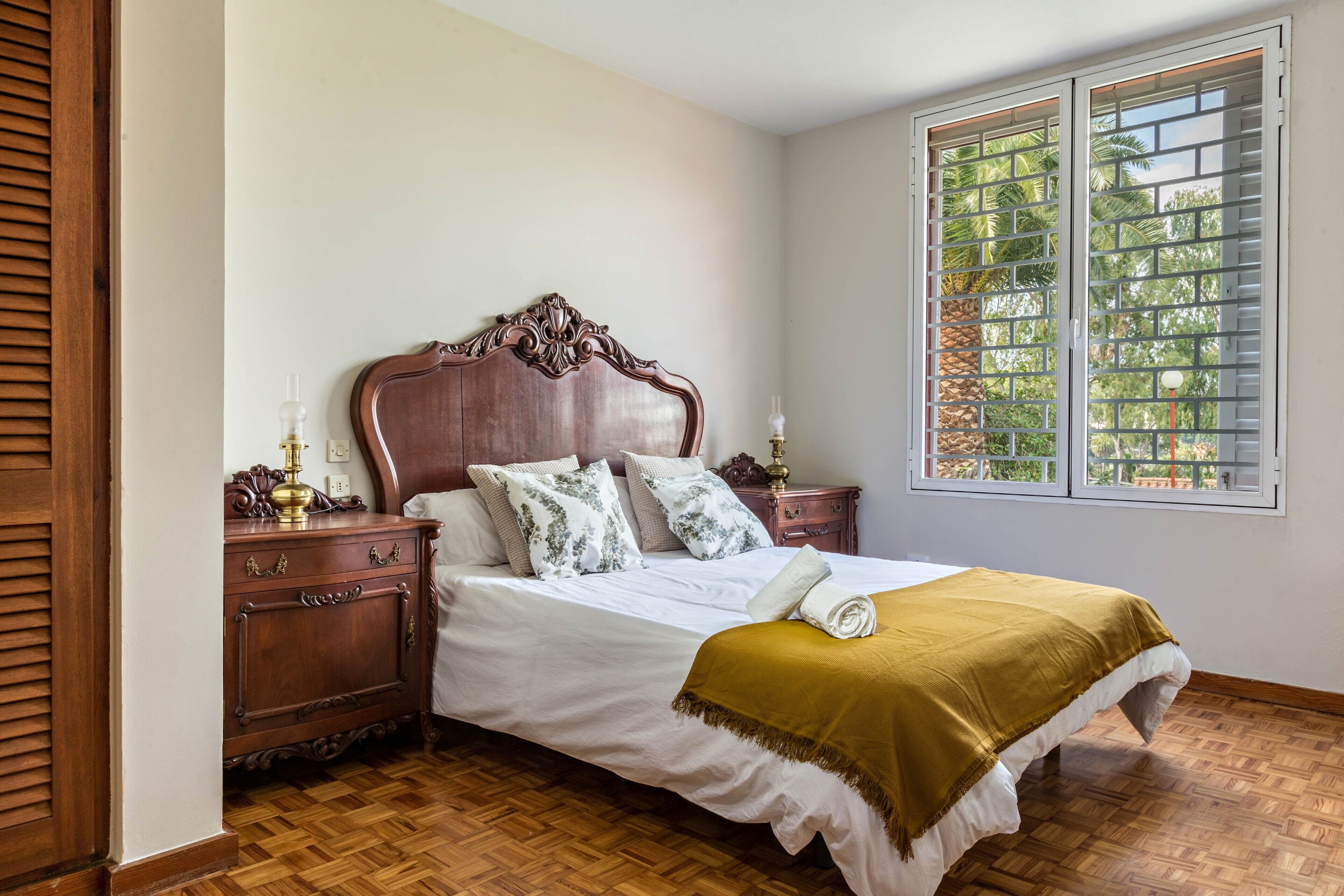 A bedroom with a wooden headboard, a bed with white bedding, a wooden nightstand, a window with blinds, and a view of greenery outside.