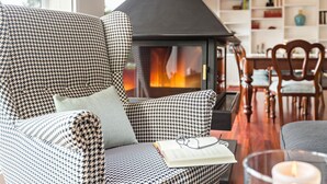 Un canapé à motifs pied-de-poule, avec un livre et des lunettes posés dessus, un poêle à bois noir et un coin repas avec des chaises en bois et une table blanche.