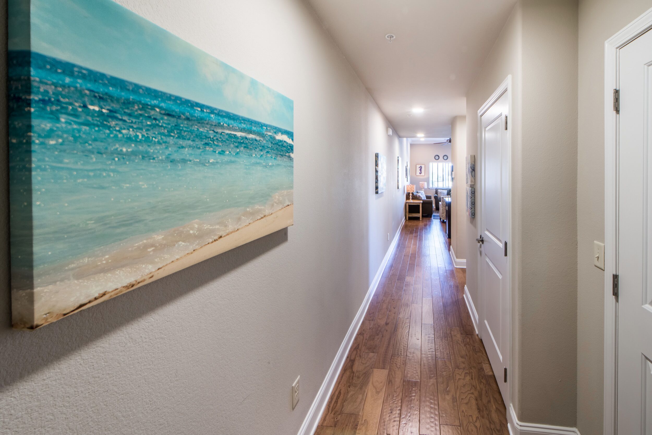 Un couloir au sol en bois, une grande peinture représentant une scène de plage et des portes blanches sur le côté droit.