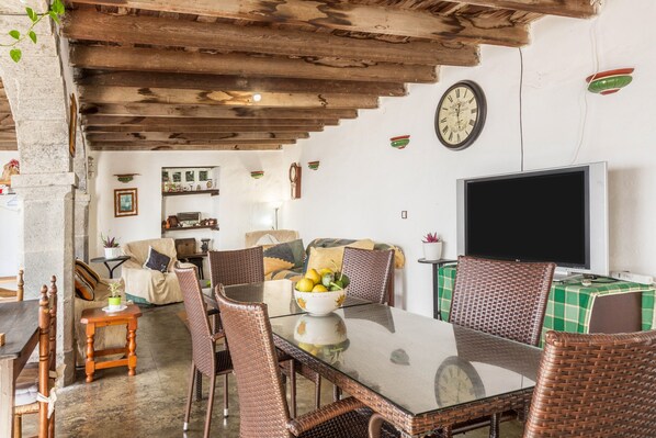 Un coin repas avec une table en verre, des chaises en osier, une horloge au mur et un téléviseur posé sur un meuble.