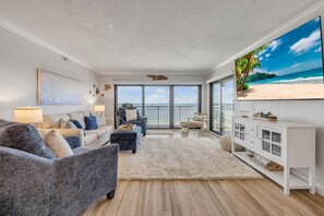 Un salon doté d’un grand téléviseur diffusant une scène de plage, d’un canapé, d’une table basse et offrant une vue sur l’océan grâce à de grandes fenêtres.
