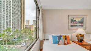 A hotel room with a large window, a bed with colorful pillows, a framed picture on the wall, and a nightstand with a lamp. - Deluxe Condo with Recreation Deck View, Free parking & WiFi (Honolulu)