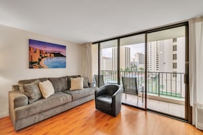 A modern living room with a grey sofa, a black leather chair, and a large window with a cityscape view. - Deluxe Condo with Recreation Deck View, Free parking & WiFi (Honolulu)