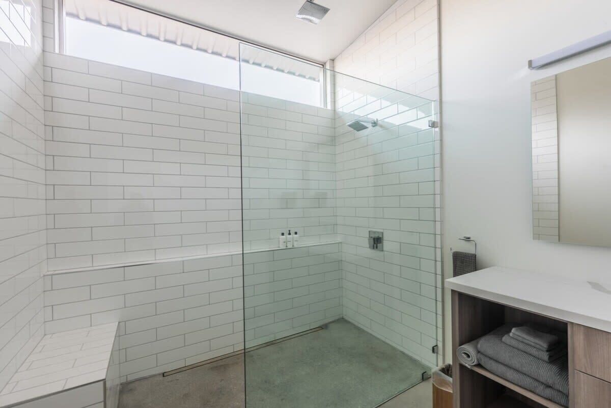 A modern bathroom with white subway tiles, a glass shower enclosure, a white sink, and a wooden shelf with towels.