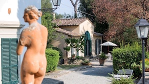 Ein Kopfsteinpflasterweg führt zu einem Gebäude mit einem Ziegeldach und grünen Fensterläden. Auf der linken Seite steht eine Statue einer Person, auf der rechten eine Laterne und im Vordergrund ein Blumentopf.