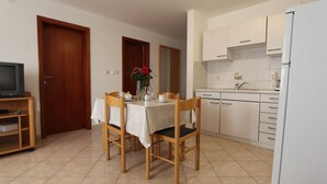 Eine kompakte Küche mit weißen Schränken, einer Spüle und einem Kühlschrank. Es gibt einen Essbereich mit zwei Holzstühlen und einem Tisch für zwei Personen. Ein Fernseher steht auf einem Holzboden links.