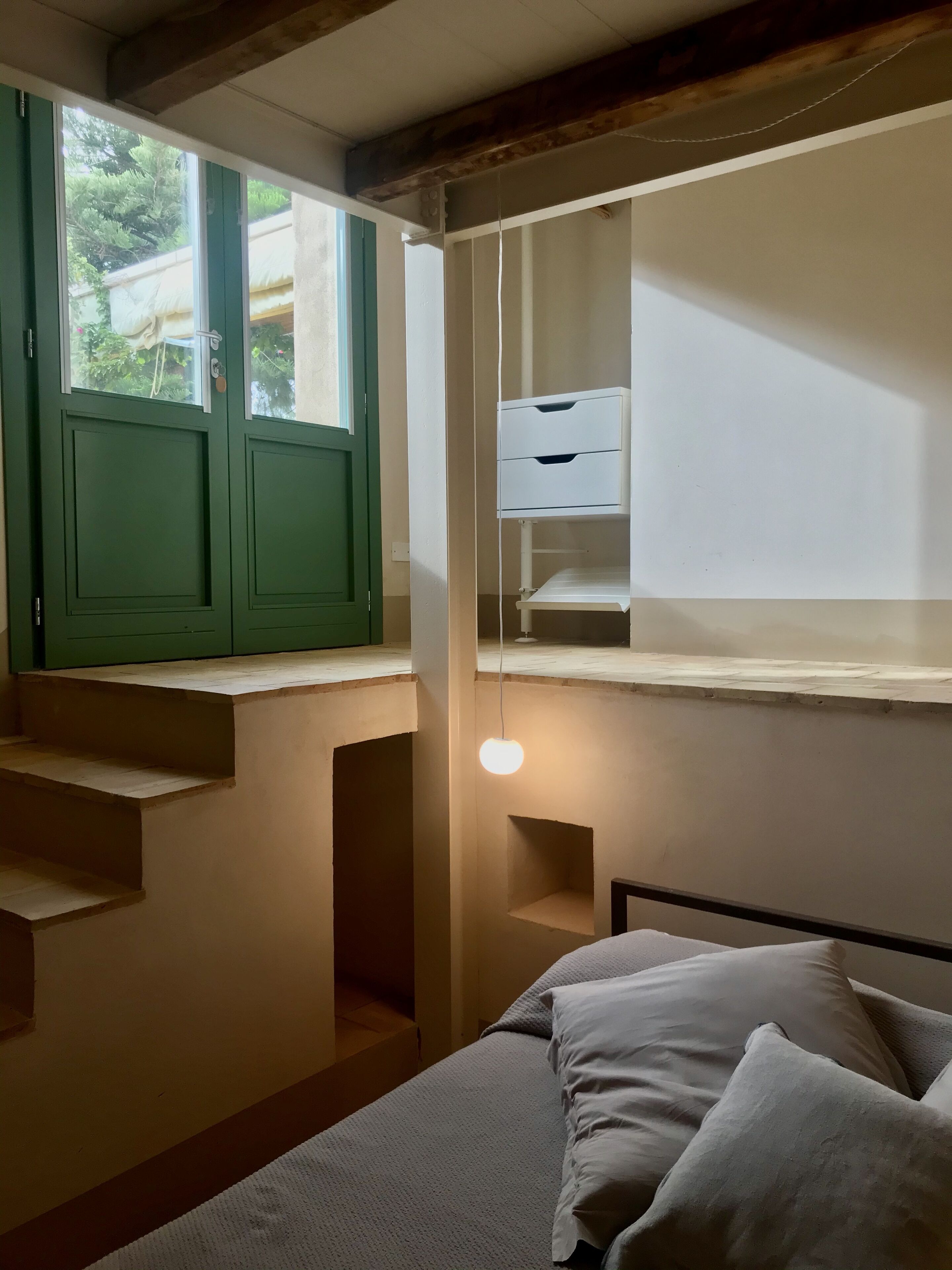 A bedroom with a bed, a window with green shutters, a desk with drawers, and a hanging light fixture.
