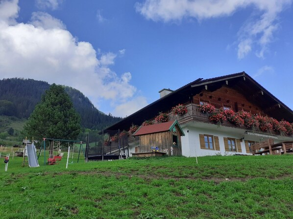 A traditional wooden chalet with a sloped roof, a balcony with red flowers, and a view of a forested mountain in the background.