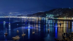 Escena costera nocturna con barcos anclados en aguas tranquilas, un paisaje urbano al fondo y un terreno montañoso.
