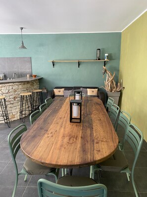 Ein Essbereich mit einem langen Holztisch, umgeben von grünen Stühlen, einer Wand aus Stein, einer Küchenzeile mit Spüle und Regalen sowie einer hängenden Leuchte.